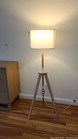 Floor lamp with wooden tripod base and white drum shade standing on a hardwood floor.