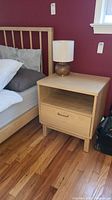 Front angled view of the oak nightstand showing the drawer closed, open shelf, and lamp on top, positioned next to a bed with a red wall background.
