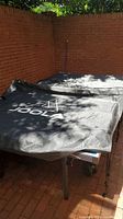 Ping pong table covered with black JOOLA cover, partially shown outdoors on brick patio.