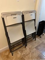 Four gray plastic Lifetime folding chairs stacked against a wall on a tiled floor, showing the chairs folded and standing upright.