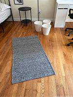 Woven gray woven throw rug laid out flat on wood floor with frayed edges visible. Three white plastic trash cans arranged together in background.