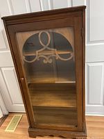 Front view of antique wooden cabinet with glass door showing decorative wood accent on glass panel and interior shelves.