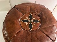 Top view showing the central floral embroidered design with stitched diamond patterns and stud accents on a round light brown leather pouffe.