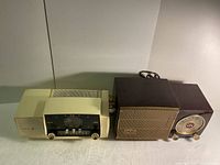 Two vintage General Electric radios side by side, showing left and front faces, cream and dark brown colors.