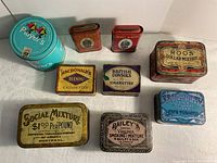 Nine vintage tobacco and cigarette tins arranged on a white surface, various brands visible including Player's, Prince Albert, MacDonald's Blends, British Consols, Roos Dollar Mixture, Edgeworth, Social Mixture, Bailey's.
