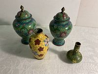 Photo showing all four items: two large lidded urns with colorful cloisonné floral designs and two smaller cloisonné vases in yellow and green, displayed on a white surface.