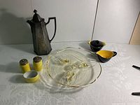 Wide shot showing all items on white cloth with subtle fern pattern: metal coffee pot, divided glass plate, yellow and black cream and sugar set, yellow shakers, small white dish