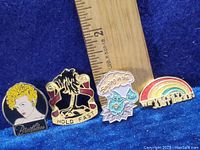 Four enamel pins on purple felt with wooden ruler for scale showing size approximately 1 to 2 inches each