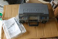 Closed gray plastic Dremel carrying case with Dremel logo on lid, placed on cardboard box, with owner's manual in plastic bag alongside.