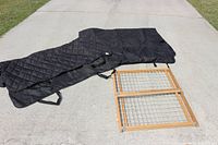 Two black quilted dog car seat covers laid flat on the ground next to a wooden dog gate with wire mesh panels.