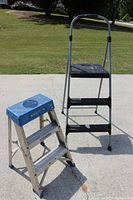 Pair of step ladders on concrete with grass background, one is aluminum with blue top, the other is taller with black steps and handle.
