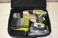 Zippered Ryobi carrying case opened showing cordless drill with battery, battery charger in netted compartment, drill bit set, and packaging inside.
