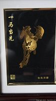 Close-up of gold foil relief sculpture of a horse with Chinese characters on black background in brown frame.
