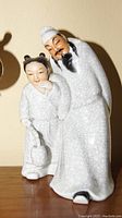 Porcelain sculpture of a Chinese man with a long beard standing next to a child with double bun hairstyle, both in traditional clothing with crackle glaze finish.