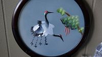 Front embroidered side showing two cranes and foliage on a blue fabric background in a round dark wooden frame.