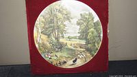 Round ceramic picture showing pastoral scene with boy, collie dog, sheep and trees; red background for display.