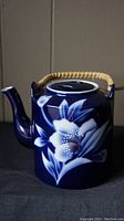 Side view of the porcelain teapot showing dark blue glaze with white and light blue floral pattern and woven rattan handle.