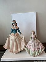 Both Coalport porcelain figurines shown together, one larger with a teal and peach dress and hat, the other smaller in green and pink dress.