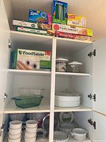 Kitchen cabinet containing various Corningware glass dishes, white ceramic quiche pans with lids, ramekins, and several types of food storage products including Ziploc and Glad bags, parchment paper, cling wrap, and FoodSaver roll.