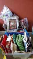 Plastic bin filled with various folded fabrics including Christmas-themed quilt pattern with Santa Claus in original packaging and assorted fabric pieces in multiple colors and patterns.