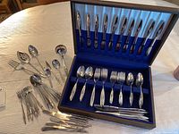 Full view of the open wooden box showing stainless steel flatware including knives, forks, spoons with additional serving utensils beside the box.