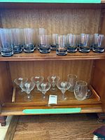 Shelf with 12 tall water or highball glasses with black bottoms, 6 cocktail or champagne glasses, and one clear coffee cup with handle.