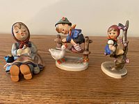 Three Goebel porcelain figurines on table: seated girl with bird, boy with goose on gate, girl by apple tree