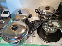 Overview of five cookware items including a large stainless steel pot with glass lid, tiered steamer pot, non-stick pans, and small pot with glass lid.