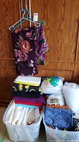 Two plastic bins filled with folded fabrics and batting, two printed cotton fabrics hanging on hangers.