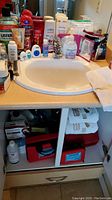 Photo showing bathroom sink, countertop with personal care items including body wash, shampoo, mouthwash, baby powder, dental floss, and a towel, plus cabinet below with more products and towels