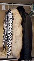 Three women's jackets hanging on hangers: black and white circular pattern cardigan, fox fur jacket, and black Helly Hansen jacket.