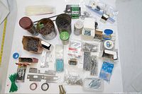 Top-down view of various nails, screws, staples, washers, metal brackets, zip ties, and rolls of tape arranged on a surface.