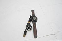 Front view of the Bushnell Neo iON golf GPS watch with black and orange perforated band and USB charger cable placed beside it on flat white surface.