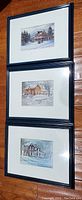 Photo showing three framed artwork prints stacked vertically on a wooden floor, each with a dark blue frame and white mat, depicting winter houses.