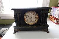 Front view of the antique mantel clock, showing black detailed casing, clock face with numbers and brass bezel.