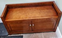 Front view of wooden storage bench showing flat seating surface with raised sides and back and two closed front cabinet doors with silver knobs.