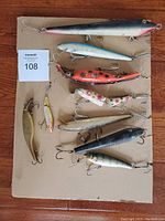 Top view of 10 assorted used fishing lures on cardboard showing various sizes, colors, and attached treble hooks.