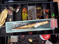 View of top tray inside the tackle box showing various metal fishing lures including spinnerbaits, spoons, small jigs, and a Rapala floating lure in original box