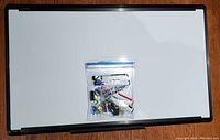 Magnetic whiteboard with black frame, 30" by 18", on wooden surface with clear plastic bag containing markers and magnets placed on board center.
