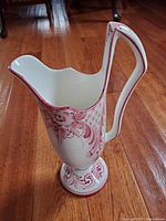 Photo of ceramic pitcher with red floral and scroll design on wooden floor.