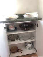 Shelved view showing various white milk glass mixing bowls with floral pattern, clear rectangular baking dishes, amber glass sauté pans, brown ceramic serving bowl with floral decorations, and stack of patterned plates.