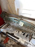 Full side view of the antique cast metal shingle cutter showing long base and lever arm, resting on a newspaper-covered surface with a note attached.