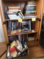 Shows a wooden cabinet with multiple shelves filled with stacked books. There is a box on the floor filled with additional books.