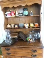 Full view of wooden hutch with items including framed prints, mugs, jars, kitchen scale, glassware and cake plate with dome.