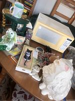 Overview of lot items displayed on table including ceramic cat figurine, plush toy, coffee cups, microwave (not included), cat framed picture, pendulum clock and decorative straw hat.