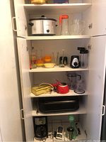 Photo of kitchen cabinet shelves containing small kitchen appliances and glassware including a white Hamilton Beach crockpot, orange and clear glass pitchers, blender parts, coffee maker, retro yellow mixer, manual juicer, salt and pepper set, and a roasting pan.