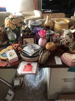 Photo shows multiple decorative plates stacked on a table along with a Franciscan pitcher with floral design, several faux flower arrangements, multiple trinket boxes including heart-shaped and oval ones, a small framed decorative item, and a light up flower vase with cord.