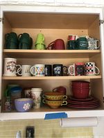 Wood cabinet shelf with multiple mugs in green, yellow, red, and multicolor including Tom and Jerry, Coca-Cola, and Busch Gardens themed mugs; bottom shelf with red plates and yellow-and-green bowls with decorative accents.