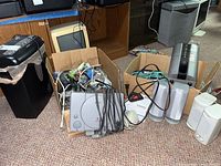 Box containing a Sony PlayStation console, wired controllers, and various video games, next to a black and gray paper shredder and some silver-gray computer speakers on the floor.