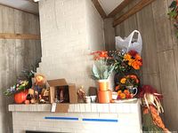 Full view of assorted seasonal decorations on mantle including ceramic pumpkins, faux flowers in pots, Halloween cup, pine cones, and scarecrow doll.